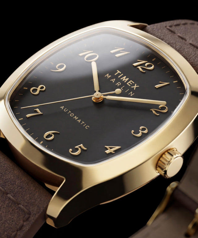 Nahaufnahme der goldfarbenen Timex Marlin Chatham Automatic mit schwarzem Zifferblatt, applizierten goldfarbenen arabischen Ziffern, goldfarbenen Zeigern und braunem Lederarmband.