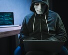 Eine maskierte, vermummte Person arbeitet in einem abgedunkelten Raum am Laptop