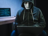 Eine maskierte, vermummte Person arbeitet in einem abgedunkelten Raum am Laptop
