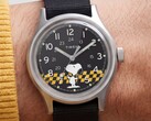 Nahaufnahme des schwarzen Zifferblatts der Timex MK1 mit Snoopy vor einer gelb-schwarz karierten Grafik sowie arabischen Stundenmarkierungen und Leuchtzeigern.