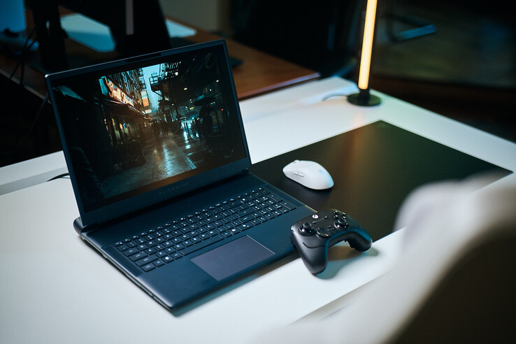 Ein aufgeklappter Alienware-Gaming-Laptop auf einem weißen Schreibtisch in einem Studio zeigt eine dunkle Straßenszene mit Reflexionen und scharfen Pathtracing-Details. Daneben liegen ein schwarzer Controller und eine weiße kabellose Maus