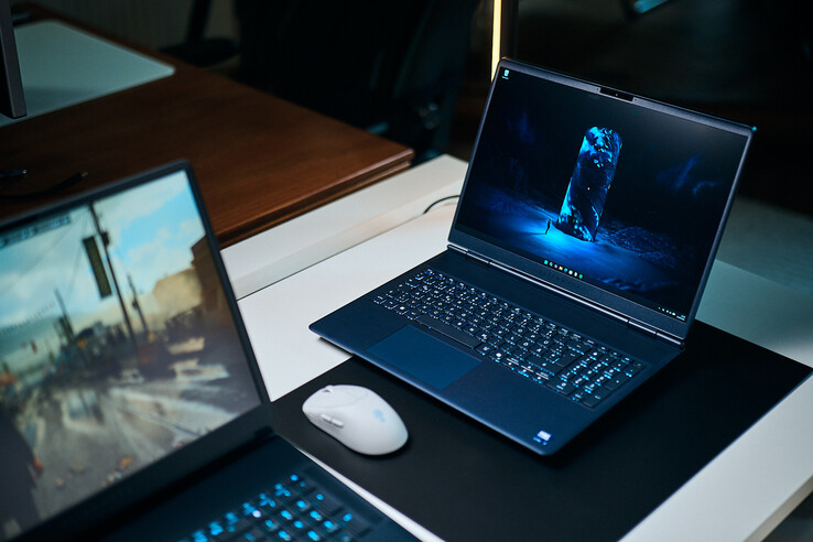 Ein dunkelblauer Mittelklasse-Gaming-Laptop (Alienware Aurora 16X) steht schräg auf einer Schreibtischunterlage und zeigt ein blau leuchtendes Science-Fiction-Hintergrundbild. Daneben liegt eine weiße kabellose Maus, während im Vordergrund ein weiterer Laptop unscharf zu sehen ist