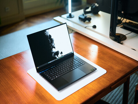 Aufgeklapptes matt-schwarzes Razer Blade 16 Gaming-Notebook auf einem Holzschreibtisch mit hellem Display-Wallpaper und einem Studio-Setup im Hintergrund.