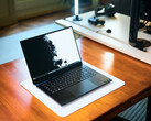 Aufgeklapptes matt-schwarzes Razer Blade 16 Gaming-Notebook auf einem Holzschreibtisch mit hellem Display-Wallpaper und einem Studio-Setup im Hintergrund.