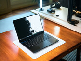 Aufgeklapptes matt-schwarzes Razer Blade 16 Gaming-Notebook auf einem Holzschreibtisch mit hellem Display-Wallpaper und einem Studio-Setup im Hintergrund.