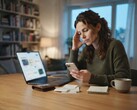 Frau am Schreibtisch schaut auf ihr Smartphone, daneben Laptop, Belege und Notizblock