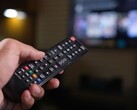 Hand hält eine Fernbedienung, im Hintergrund ein unscharfer Fernseher