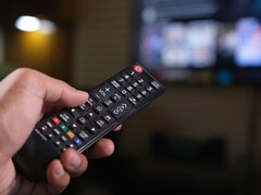 Hand hält eine Fernbedienung, im Hintergrund ein unscharfer Fernseher