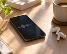 Smartphone mit Audio-Wellenform auf dem Display neben kabellosen Ohrhörern und einer Kaffeetasse auf einem Holzschreibtisch