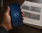Hand hält ein Smartphone mit Chatbot-Symbol, daneben liegt eine gedruckte Zeitung.