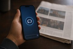 Hand hält ein Smartphone mit Chatbot-Symbol, daneben liegt eine gedruckte Zeitung.