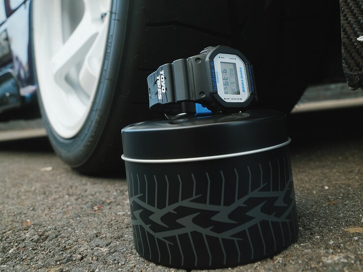 Eine Casio G‑Shock DW5600TT25‑1 x Toyo Tires wird auf einer metallenen Dose mit Reifenmotiv präsentiert, direkt neben einem Autorad