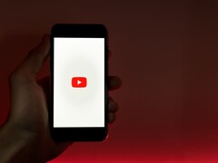Hand hält ein Smartphone, auf dem Display leuchtet das YouTube-Logo vor rotem Hintergrund