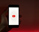 Hand hält ein Smartphone, auf dem Display leuchtet das YouTube-Logo vor rotem Hintergrund