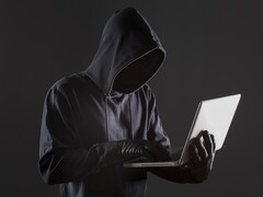 Ein vermummter Hacker arbeitet an einem Laptop vor dunklem, anonymem Hintergrund
