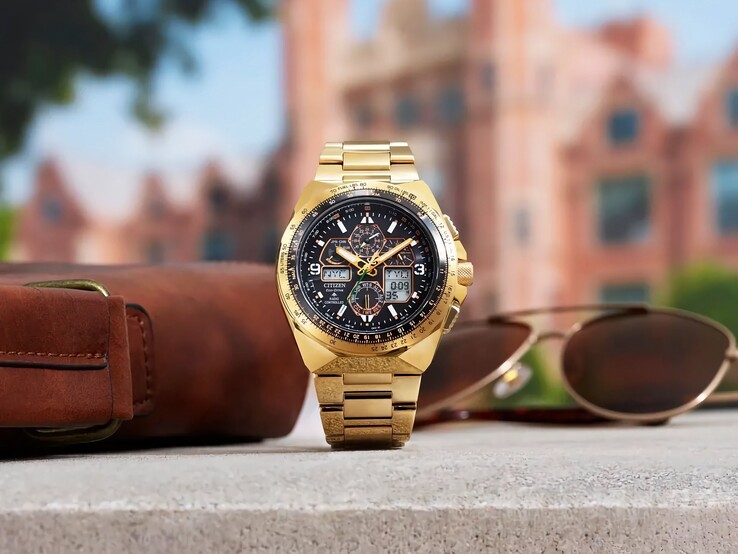 Goldfarbene Fliegeruhr Citizen Promaster Skyhawk A-T JY8144-50E, aufrecht auf einem Absatz neben einer braunen Ledertasche und einer Sonnenbrille, mit unscharfer Stadtkulisse im Hintergrund