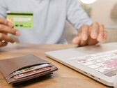 Person hält eine Bankkarte vor einem Laptop, daneben liegt ein Portemonnaie