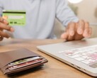 Person hält eine Bankkarte vor einem Laptop, daneben liegt ein Portemonnaie