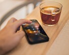 Hand tippt eine PIN auf einem Smartphone, das neben einem Glas Tee auf einem Holztisch liegt