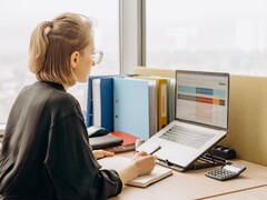 Frau mit Brille arbeitet am Laptop an einer Tabellenkalkulation, daneben Ordner und ein Taschenrechner