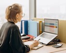 Frau mit Brille arbeitet am Laptop an einer Tabellenkalkulation, daneben Ordner und ein Taschenrechner