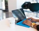 Hände tippen auf einem Laptop mit blauer Tastatur, daneben ein Becher mit Microsoft-Logo