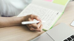 Person schreibt Notizen in ein Notizbuch neben einem aufgeklappten Laptop