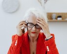 Ältere Frau mit weißem Haar und Brille telefoniert in einem hellen Wohnraum und fasst sich besorgt an die Schläfe.