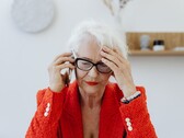 Ältere Frau mit weißem Haar und Brille telefoniert in einem hellen Wohnraum und fasst sich besorgt an die Schläfe.