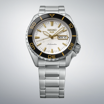 Seiko 5 Sports SKX HDB004 mit silberweißem Zifferblatt, schwarzer Lünette mit goldfarbenen Markierungen, goldfarbenen Zeigern und Indizes sowie Edelstahlarmband vor hellgrauem Hintergrund